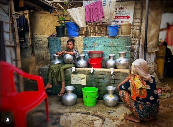 Girl sitting on a brick wall, surrounded by pots of drinking water, with a woman covering her hair squatting in the foreground on the right