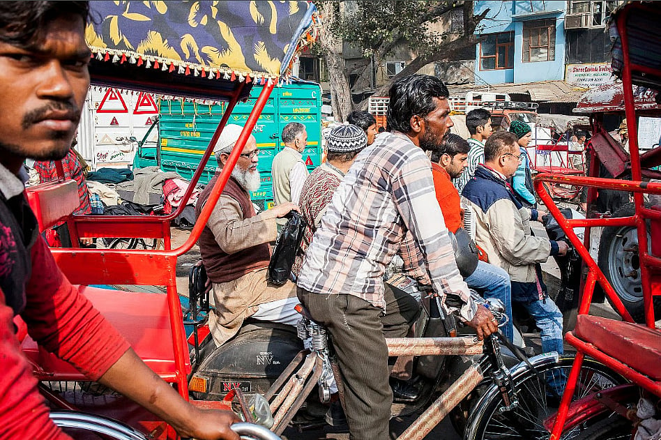 Photo of people on bicycles and red rickshaws crowding a street in Old Delhi. On the left a man driving a rickshaw is looking into the camera. The others are looking ahead, intent on where they are going.