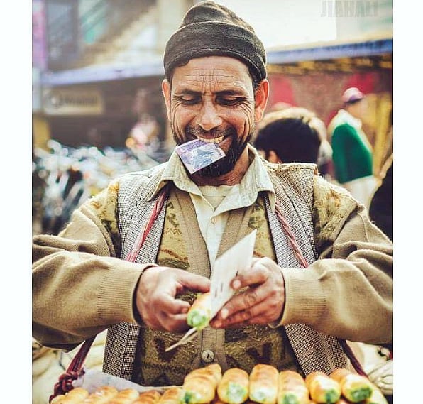 Photo of a corn seller wearing a beanie hat. He is smiling as he wraps an ear of corn to sell and is holding a note of currency in his teeth.