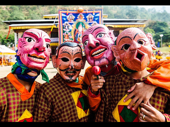 Photo of masked dancers. The masks are old and wooden, and there are four dancers, wearing a variety of faces. They are also wearing checked shirts. 