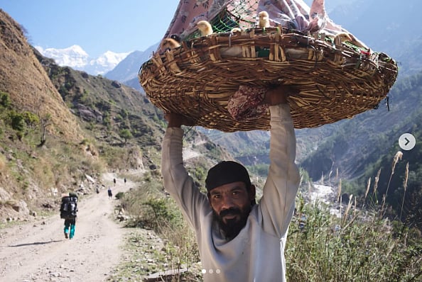 Man holding cane basket of produce. He is standing on a hiking trail and a hiker can be seen in the distance. He is surrounded by mountains.