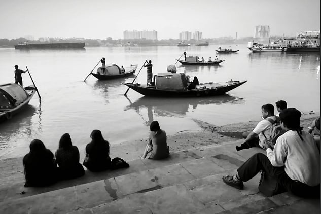 Photo of women sitting on the banks of a river in hijab, looking out at boats which are being rowed on the river. The photo is in black and white 