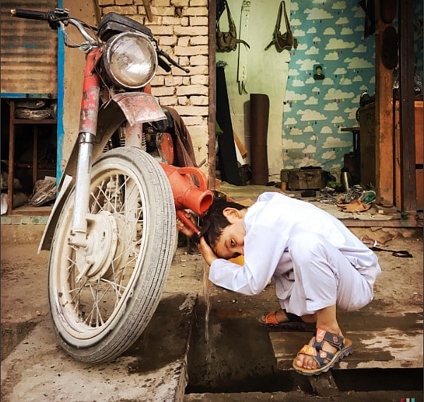 Boy washing his hair from a plastic kettle over a drain and next to a bike. 