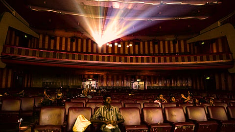 A wide shot of a dimly lit, vintage cinema hall with worn-out red seats. A few scattered viewers sit in the rows, looking up toward a bright beam of light shining from the projection booth.