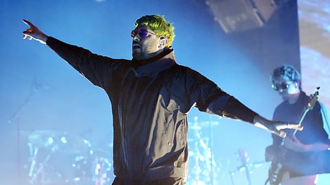 Jai Paul performing live on stage with tinted glasses, arms outstretched under blue stage lighting.