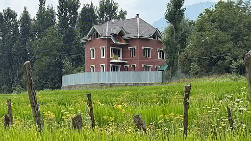 A new brick house, protected by a fence, rises from the middle of a field, a powerful symbol of farmland lost to construction.