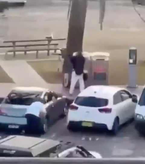 One of the gunmen (dressed in dark clothing) holding a long rifle/shotgun. A bystander, Ahmed El Ahmad (wearing a light-colored shirt), approaches  crouching behind parked cars.