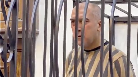 Atef Najib, Bashar al-Assad's cousin and former Daraa security chief, standing in a metal cage wearing a striped prison uniform.