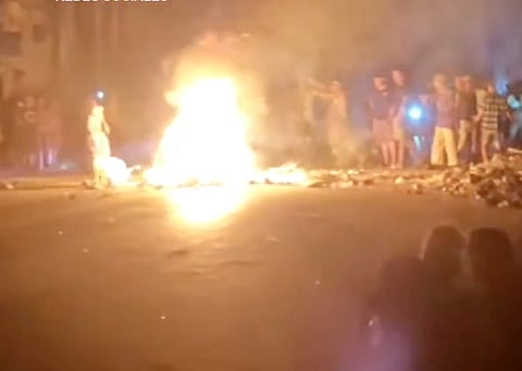 Nighttime crowds in Marianao, Havana blocking streets, banging pots, and burning trash piles in protest after over 20 hours without power, amid thick smoke and debris.