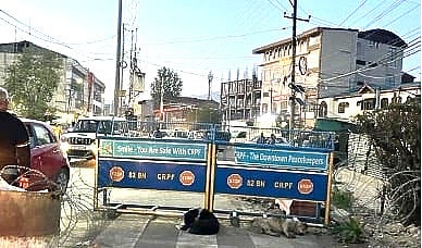 A street barricade fit with barbed wire blocks two-way flow of traffic. It reads, “Smile you are safe with CRPF (Central Reserve Police Force) & CRPF - The Downtown Peacekeepers.” CRPF is an Indian federal armed police force. It operates extensively in Kashmir under the mandate of internal security and leading counter-insurgency efforts in the region.