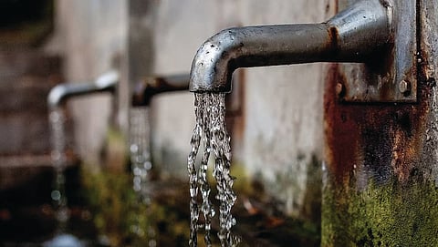 A close-up, low-angle shot of water streaming from a row of weathered metal outdoor taps attached to a mossy wall.