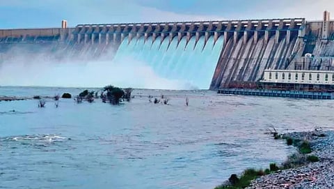 A massive concrete dam with dozens of open floodgates releasing powerful, turbulent white water into a broad riverbed under a clear blue sky.