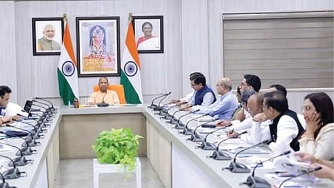 A wide-angle shot of a formal meeting room where CM Yogi Adityanath is seated at the head of a U-shaped table, flanked by national flags and portraits, with several officials seated along the sides.