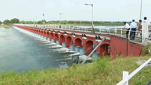 A long, red-arched reservoir structure with multiple open gates through which water is being discharged into a large basin, with officials observing from a walkway above.