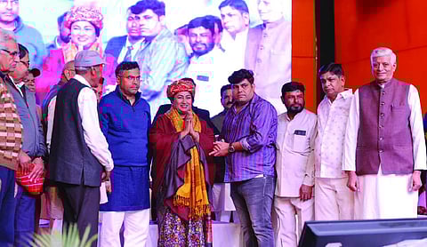 Chief Minister Rekha Gupta, wearing a traditional colorful turban and a red shawl, stands with a group of officials on a stage during the foundation stone ceremony. A large screen in the background displays event branding as she greets the crowd with a 'Namaste'.