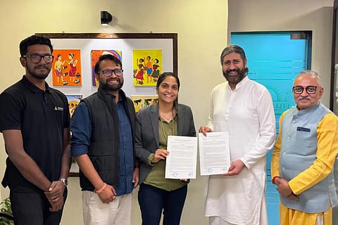 A group of five people stands in a modern office setting. In the center, a woman in a grey blazer and a man in a white long tunic hold up signed copies of a Memorandum of Understanding (MoU). They are flanked by three men in professional and traditional attire. On the wall behind them are four colorful, traditional Indian paintings, and a frosted glass door with a sun logo is visible to the right.