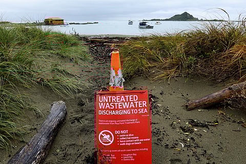 An expansive aerial photograph showing the Moa Point Wastewater Plant nestled between the Wellington Airport runway and the rugged South Coast. A coastal road winds past residential houses toward the airport, with the grey Cook Strait waters meeting a rocky shoreline under a cloudy sky.