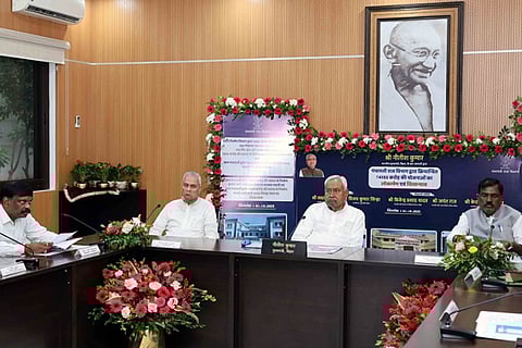 Bihar Chief Minister Nitish Kumar and senior cabinet ministers sit at a long dark-wood conference table during an official meeting. The room is decorated with vibrant pink and red flower arrangements, and a large framed portrait of Mahatma Gandhi hangs on the wall behind the CM. A backdrop features official government branding in Hindi regarding urban development schemes.