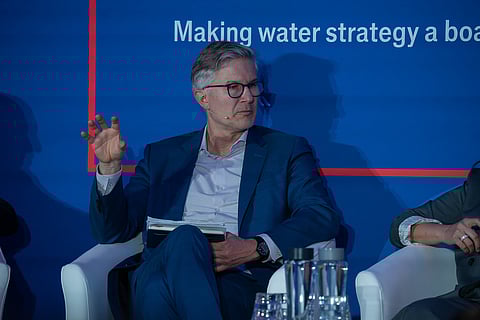 A medium shot of Dr. Helge Daebel, a man with grey hair and glasses, wearing a sharp blue suit and a light-colored shirt. He is seated on a white armchair and gesturing with his right hand during a panel discussion. Behind him is a deep blue wall featuring the white text 