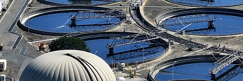 An aerial view of several large, circular primary clarifier tanks at a wastewater treatment plant, featuring rotating bridges and a large white anaerobic digester dome in the foreground.