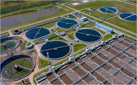 High-angle shot of an industrial sewage treatment plant featuring multiple blue circular primary clarifiers and a grid of brown rectangular aeration tanks surrounded by green landscape.