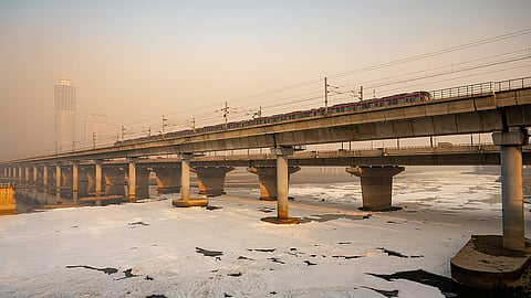 A thick layer of white pollution foam covering the surface of the Yamuna River beneath a multi-tiered metro and traffic bridge under a hazy sky.