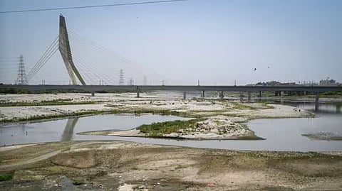 The dried-up, shallow riverbed of the Yamuna River in Delhi with sand patches exposed, featuring the Signature Bridge in the background under a clear sky.