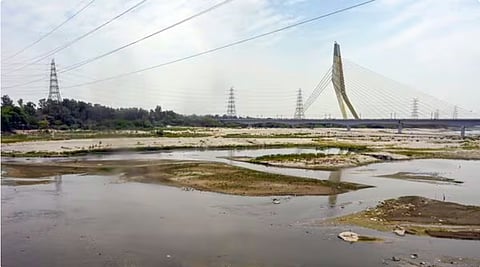 A wide shot of a drying river landscape featuring small streams flowing through sandy banks, with the Signature Bridge and electric transmission towers under a hazy sky.