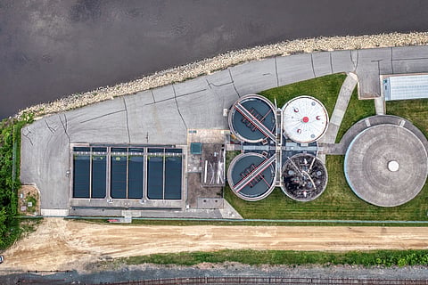 The facility includes four long rectangular pools, a cluster of four circular processing tanks, and one larger independent circular concrete structure, bordered by paved roads, green grass, and a rocky shoreline.