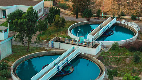 Aerial view of two large, circular concrete wastewater clarifier tanks with blue-tinted water and central bridge scrapers, surrounded by green lawns and industrial buildings.