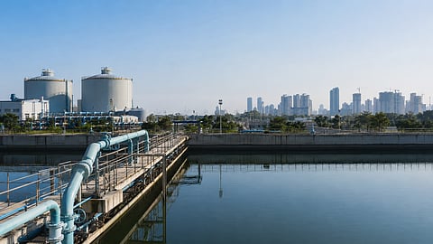 Water-treatment facility with large pipelines, storage tanks and a city skyline reflected in a calm reservoir.