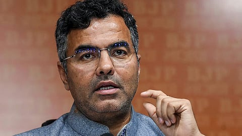 Close-up of a man wearing glasses and a grey jacket, speaking and gesturing during a media interaction.