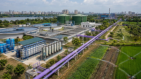 Modern wastewater treatment plant with clarification tanks, membrane filtration systems, reclaimed-water storage and purple pipelines distributing treated water to an industrial area, landscaped spaces and irrigated farmland.