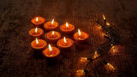 A set of beautifully hand-painted, earthy clay diyas lit with a warm, golden flame, placed on a traditional Indian wooden surface next to a single, unlit, tangled string of bright, synthetic electric lights. 
