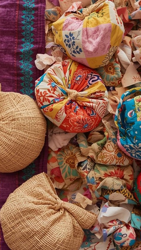 Close-up of a mix of vibrant textiles and bundles. Features two gathered pouf-like objects made from colorful floral patchwork cotton and two round bundles made from natural jute, all set against a background of purple and teal fabric and many loose fabric scraps.