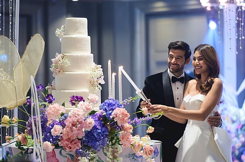 A man in black elegant suit and a woman in a white wedding dress lights the candles next to a 6-tier wedding cake decorated with beautiful purple, pink and lavender flowers.