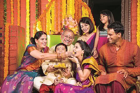 Happy Indian Family Celebrating Ganesh Festival or Chaturthi - Welcoming or performing Pooja and eating sweets in traditional wear at home decorated with Marigold Flowers