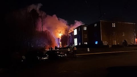Flames consume an apartment building at night, illuminating the scene with a bright orange glow amidst the darkness.