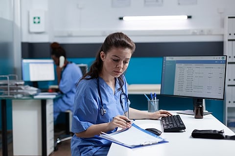 A female nurse in scrubs seated at a desk, focused on her work with medical tools and papers around her.