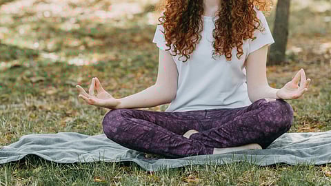 A woman sits cross-legged on the grass in a park, eyes closed, practicing meditation amidst trees and sunlight.