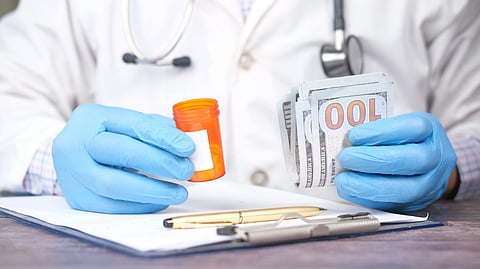 A doctor wearing blue gloves is holding a orange colored pill container in one hand and a stack of dollars.