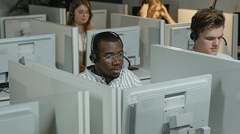 A group of call center employees wearing headsets are seated in a cubicle space, working on their computers.