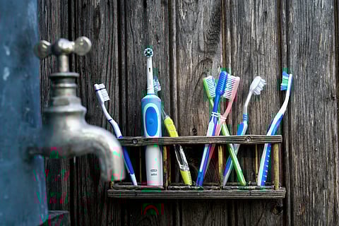 Toothbrushes, including one electric, stored in a wooden rack by a rustic wall with a metal water tap on the side.