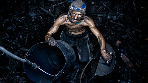 A shirtless, soot-covered coal mine worker holds metal buckets amidst dark charcoal debris.