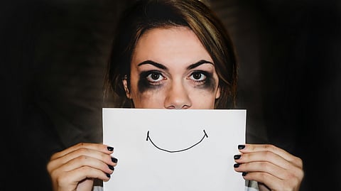 A woman with smudged eye makeup holds a paper with a drawn smile over her mouth.