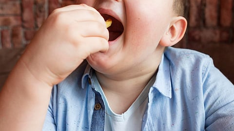 An obese kid wearing a blue shirt, sitting on a sofa, and eating French fries.