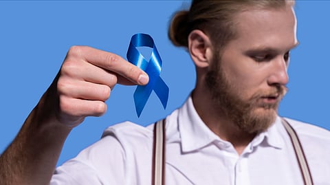 A bearded man wearing a white shirt is holding a blue prostate cancer awareness ribbon.