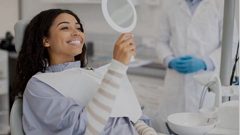 A woman is smiling and looking at herself in a mirror in hand sitting on a dental chair.