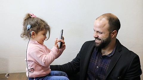 A child happily looking at mobile screen while wearing a hearing aid and her father looks happily at her.