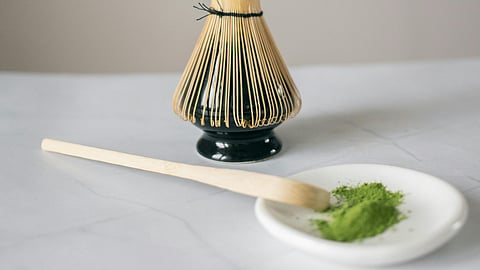 A green powder is kept on a white plate with a wooden spoon and a bamboo whisk is kept nearby.